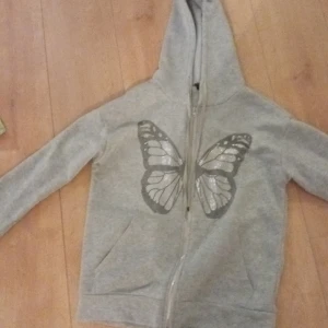 Grå hoodie med fjärilstryck - Säljer en superfin grå hoodie med en stor fjäril i silvertryck på framsidan. Den har en dragkedja och huva med snören. Perfekt för en avslappnad stil och passar till alla årstider. Skön och mysig att ha på sig!