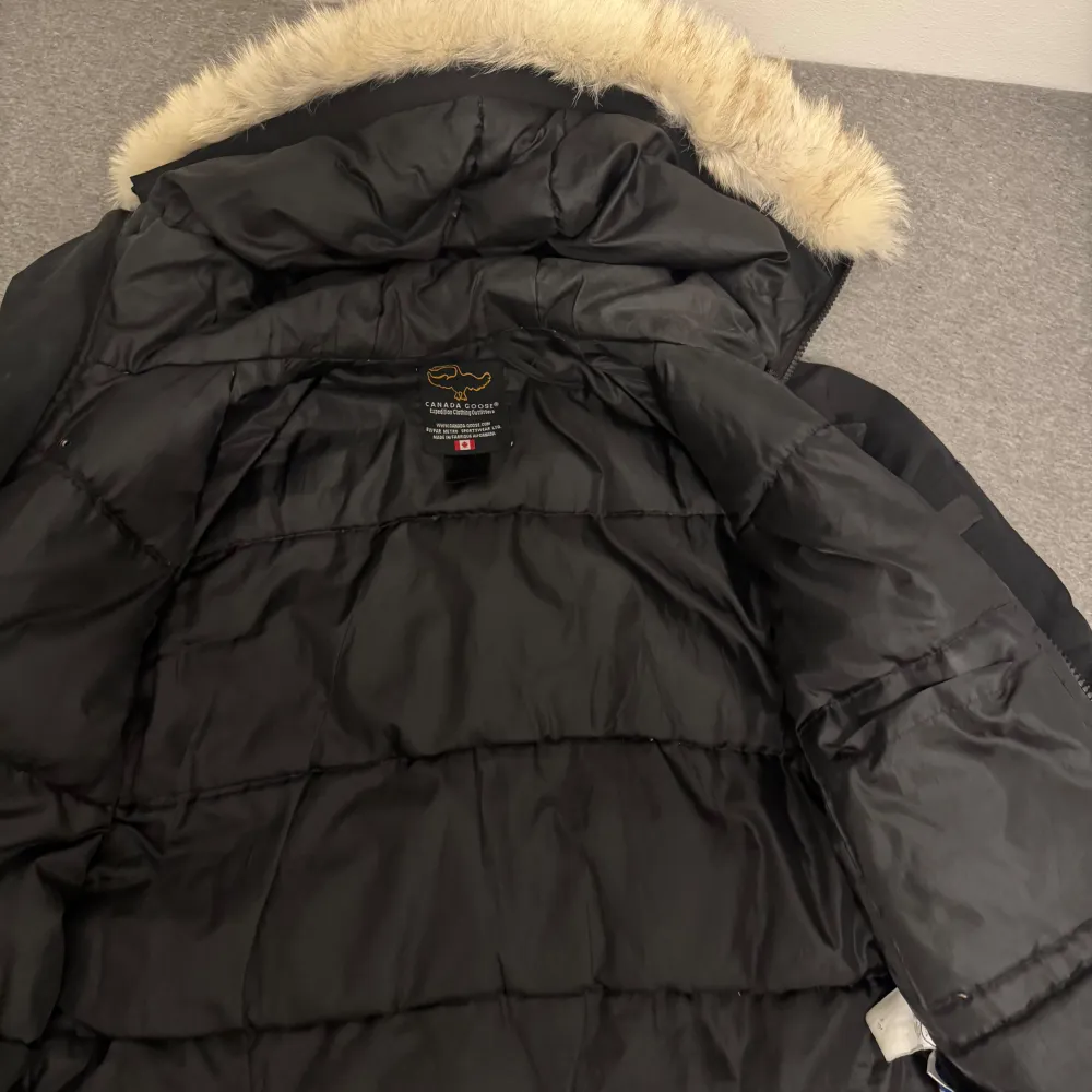 Säljer en svart dunjacka från Canada Goose med en luva med päls på. Jackan har en dragkedja och knappar framtill, samt flera praktiska fickor. Perfekt för kalla vinterdagar. . Jackor.