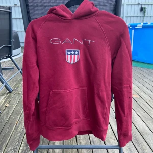 Vinröd hoodie från GANT - Säljer en snygg vinröd hoodie från GANT med broderad logga och emblem på framsidan. Den har en stor ficka fram och en bekväm huva. Perfekt för kyliga dagar eller en avslappnad stil. Passar både till vardags och chillkvällar. Aldrig använd då jag fick den i present och röd är inte min färg.