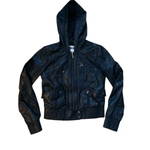 Svart skinnjacka med huva från Topshop - Säljer en snygg svart skinnjacka från Topshop i storlek 34. Jackan har en cool huva och dragkedja framtill. Den har också praktiska fickor med knappar. 