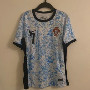 Portugal fotbollströja med Ronaldo  - Säljer en cool Portugal fotbollströja från Nike med Storlek S den passar M ochså den har nummer 7 på framsidan och 'Ronaldo' på baksidan. Tröjan har ett unikt blått mönster på vit bakgrund och svarta detaljer. Perfekt för fotbollsfans! 🏆Pris kan jättegärna diskuteras  Ps den är aldrig använt jag testa den 1 gång och den passade inte mig jag säljer den billigast av alla andra här bara skriva om du har frågor Elle sånt.