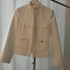 Beige kavaj med guldknappar - Snygg beige kavaj med långa ärmar och stilrena guldknappar. Perfekt för höst och vår, med en klassisk look som passar både till vardags och fest. Två fickor framtill ger en extra touch av elegans.