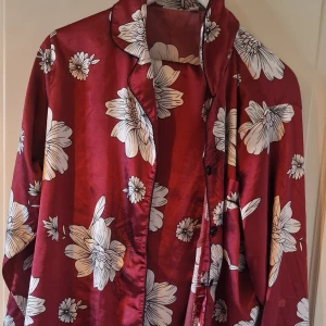 Vinröd blommig satanpyjamas  - Säljer en snygg vinröd satanpyjamas  med stora vita blommor. Satanpyjamas har långa ärmar och knappar framtill. Perfekt för en avslappnad men stilren look. Passar bra till både vardag och sova!