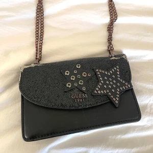 Svart axelväska från Guess - Snygg svart axelväska från Guess med coola stjärndetaljer i silver och nitar. Tyvärr är den väl använd och sliten på ovansidan och finns även tecken på användning inuti🥲