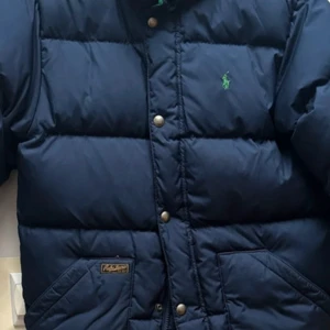 Mörkblå dunjacka från Ralph Lauren - Storlek m för barn 10-12 år!!!Säljer en snygg mörkblå dunjacka från Ralph Lauren. Jackan har en broderad grön logga på bröstet och knäppning med knappar. Perfekt för kalla dagar med sin varma och puffiga design. Passar både till vardags och lite finare tillfällen.