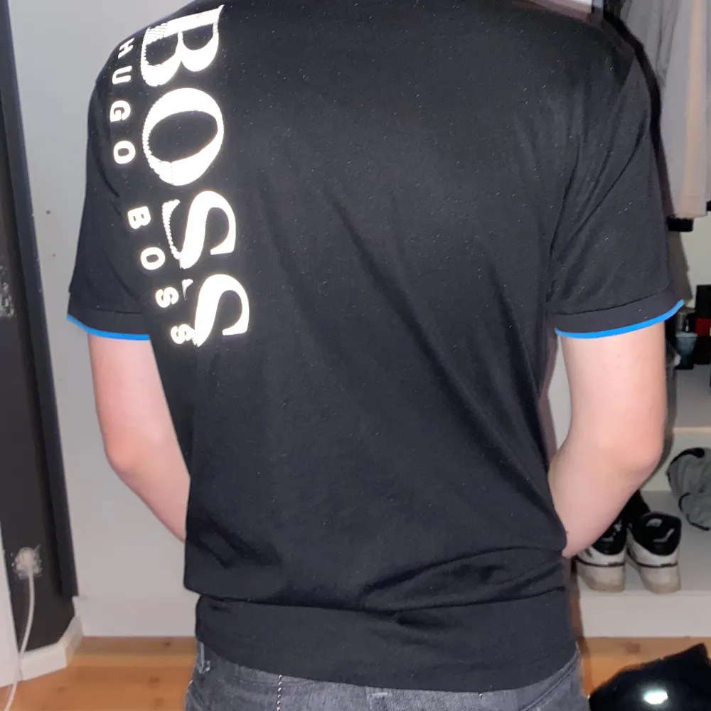 Snygg svart pikétröja från Hugo Boss i slim fit. Den har en klassisk krage med blå detaljer och knappar framtill. Perfekt för både vardag och fest. Tröjan har korta ärmar och är i mycket bra skick.. T-shirts.