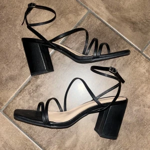 Svarta sandaler med klack - Snygga svarta sandaler med klack och smala remmar som korsar foten. Perfekta för en utekväll eller fest! De har en fyrkantig tå och justerbara spännen runt ankeln för bästa passform. Endast använda 2 kvällar, i mycket bra skick. Storlek 39, normala i storlek.