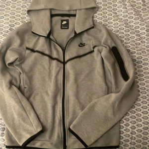 Grå Nike Tech - Säljer en grå hoodie från Nike i storlek S. Den har en dragkedja framtill och en huva för extra mysfaktor. Perfekt för både träning och chill. Den är i bra skick!