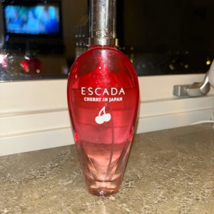 Escada Cherry in Japan Parfym - Säljer en superfin parfym från Escada, 'Cherry in Japan'. Flaskan är röd och rosa med en elegant, avsmalnande form och ett silverfärgat lock. Doften är fruktig och söt, perfekt för vår och sommar. Flaskan är nästan full, så det finns mycket kvar att njuta av! 🍒