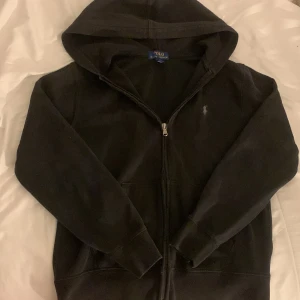 Svart hoodie från Polo Ralph Lauren - Säljer en svart hoodie från Polo Ralph Lauren i bra skick. Den har en dragkedja framtill och en klassisk logga på bröstet. Perfekt för höst och vår, med långa ärmar och en skön huva. Passar både till vardags och chillkvällar. 🖤