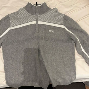 Grå tröja med dragkedja från Boss - Säljer en stilren grå tröja från Boss med dragkedja och hög krage. Tröjan har vita och ljusgrå ränder över bröstet och längs ärmarna. Perfekt för både vardag och lite finare tillfällen. Den är i mycket bra skick och superbekväm att ha på sig.