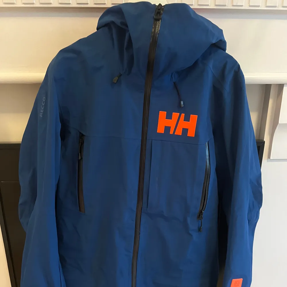 Säljer en snygg blå jacka från Helly Hansen med orange logga på bröstet. Jackan har en dragkedja framtill och är utrustad med RECCO-teknologi. Perfekt för höst- och vinteraktiviteter. Den har en justerbar huva och långa ärmar. Passar både för vardag och utomhusäventyr.. Jackor.