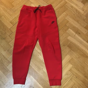 Röda mjukisbyxor från Nike - Säljer ett par snygga röda mjukisbyxor från Nike i storlek L. De har en elastisk midja med snörning och en praktisk dragkedja på baksidan. Perfekta för träning eller en chill dag hemma. Superbekväma och i bra skick!