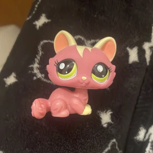 Liten rosa kattfigur från LPS  - Söt liten kattfigur från Littlest Pet Shop i rosa med stora gröna ögon och vita detaljer. Perfekt för samlare eller som en gullig dekoration. Figuren är i bra skick och har märkningen 'LPS' på sidan. En charmig liten prydnad för alla LPS-fans! Gen 3 eller 4!