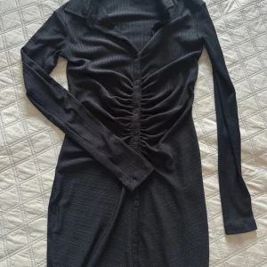 Svart ribbad klänning - Snygg svart ribbad klänning med långa ärmar och knappar framtill. Den har en smickrande rynkad detalj vid midjan som ger en fin passform. Perfekt för höst och vinter, både till vardags och festligare tillfällen. 167 cm och slutar precis strax över knäna. 
