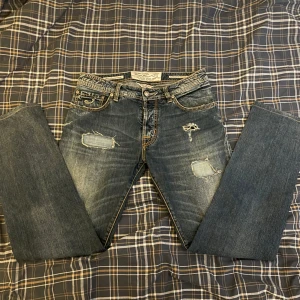 Blå/grå jeans från Jacob Cohen - Säljer ett par snygga blå/grå jeans från Jacob Cohen i storlek 33. De har en cool slitning på framsidan och knappar istället för dragkedja. Perfekta för en avslappnad stil. Passar bra till både vardag och fest!