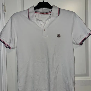 Vit pikétröja från Moncler - Säljer en snygg vit pikétröja från Moncler i storlek S. Tröjan har korta ärmar och en klassisk krage med röd och blå randig detalj. Perfekt för både vardag och fest! 🕺