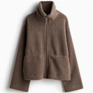 Brun teddyjacka med dragkedja - Mysig brun teddyjacka perfekt för kyliga dagar. Den har en dragkedja framtill och stora fickor för extra värme och stil. Jackan är långärmad och har en hög krage som skyddar mot vind. Perfekt för höst och vinter!