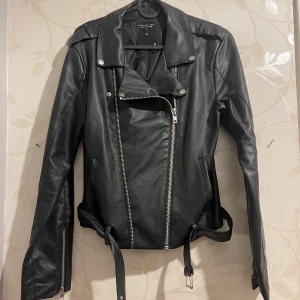 Svart skinnjacka med dragkedja - Snygg svart skinnjacka i mycket bra skick. Den har en cool asymmetrisk dragkedja och bälte i midjan för extra stilpoäng. Perfekt för höst och vår, med långa ärmar och en klassisk biker-look. Passar både till vardags och fest!