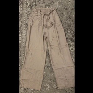 Beige byxor med knytband - Säljer ett par snygga beige byxor med hög midja och knytband i midjan. De har en loose passform och vida ben, vilket gör dem superbekväma och perfekta för både vardag och fest. Materialet känns lätt och luftigt, perfekt för vår och sommar. Passar bra till en enkel topp eller en mer dressad blus. Det är bara att stryka den så blir den som ny igen 