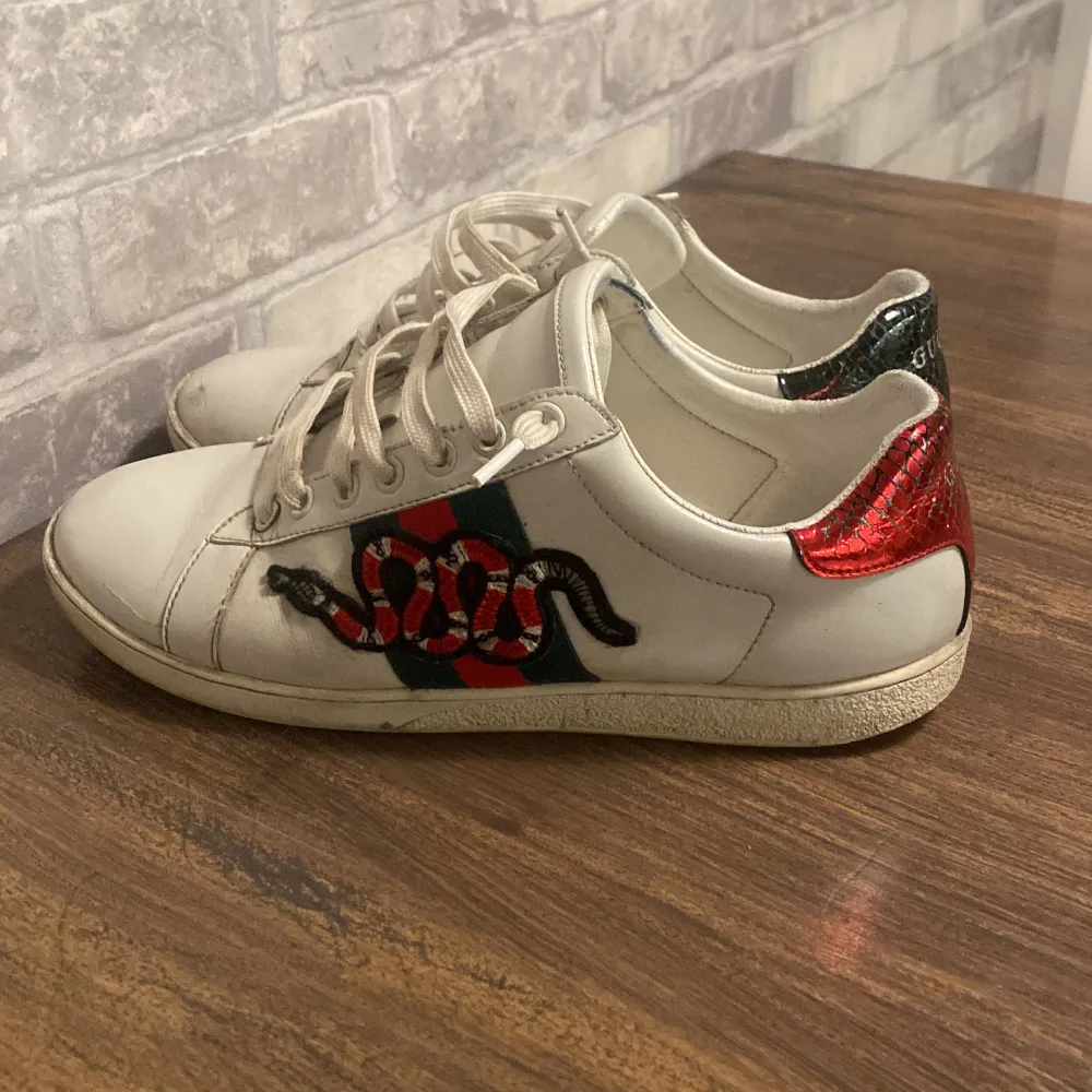 Säljer ett par vita Gucci-sneakers i skinn med ikoniska ränder i grönt och rött. Skorna har en cool ormmönsterdetalj på sidan och glittriga hälpartier i rött och grönt. Perfekta för att ge din outfit en lyxig touch! Använda men i bra skick.. Skor.