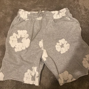 Grå shorts med blommönster från Denim Tears - Säljer ett par grå shorts från Denim Tears i storlek M. De har ett coolt vitt blommönster och snörning i midjan för perfekt passform. Perfekta för en avslappnad sommarstil! 🌼