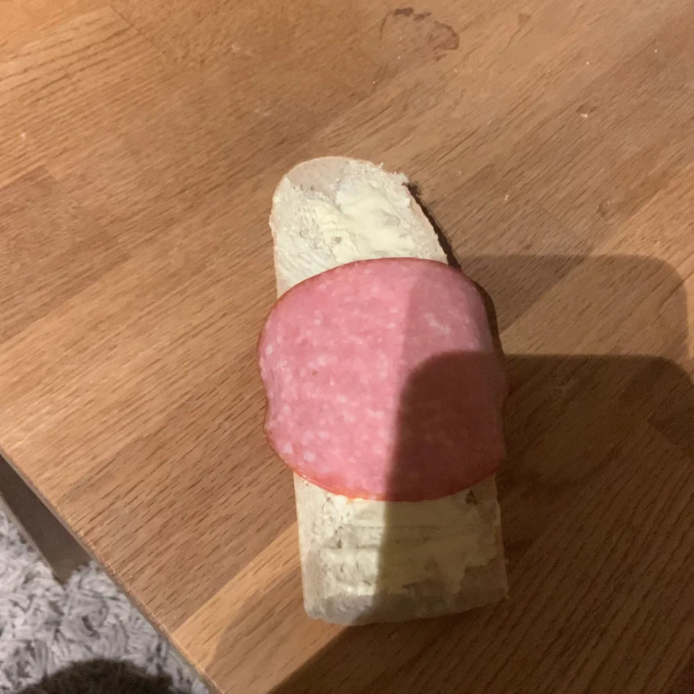 En skiva baguette med smör och en skiva salami ovanpÄ, placerad pÄ ett trÀbord. Priset kan diskuteras .. Asusteet.
