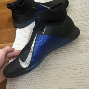 Svart och blå Nike fotbollsskor - Säljer ett par svarta och blå Nike fotbollsskor i mycket bra skick. Skorna har en snygg design med silverdetaljer och är perfekta för inomhusfotboll. De har en tight passform och är gjorda i syntetmaterial för bästa hållbarhet och prestanda.
