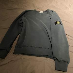 Säljer en snygg grå sweatshirt från Stone Island i mycket bra skick. Tröjan har en klassisk rund halsringning och den ikoniska loggan på ärmen. Perfekt för både vardag och chill. Passar bra till jeans eller joggers. Skön och stilren design!