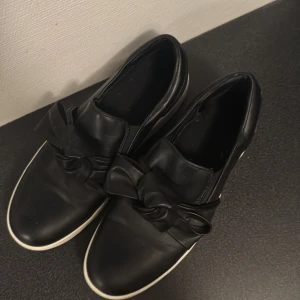 Svarta sneakers med rosett - Säljer ett par snygga svarta sneakers i skinn med en cool rosettdetalj på ovansidan. De har en vit sula som ger en fräsch kontrast och är perfekta för både vardag och fest. Skorna är i bra skick och passar till många olika outfits. Perfekta för dig som vill ha något stilrent men ändå med en twist!