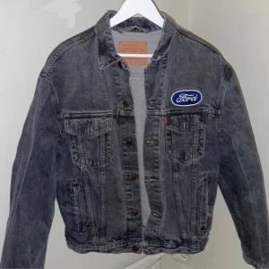 Grå jeansjacka från Levi's med Ford-märke - Säljer en grå jeansjacka från Levi's i bra skick. Den har ett coolt Ford-märke på bröstet och klassiska bruna knappar. Perfekt för höst och vår, med en avslappnad passform. Jackan är långärmad och har två bröstfickor med lock. Perfekt för dig som gillar en retro look!
