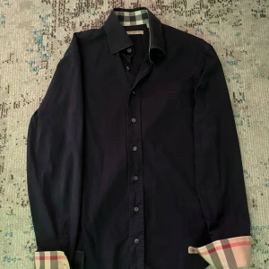 Svart skjorta från Burberry - Snygg svart skjorta från Burberry i bomull. Den har långa ärmar och klassiska knappar framtill. Insidan av kragen och ärmsluten har det ikoniska Burberry-rutiga mönstret i beige, rött och svart. Perfekt för både vardag och fest!