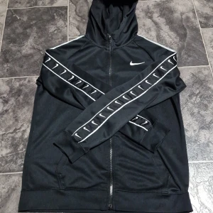 Nike huvtröja - Säljer en svart huvtröja från Nike i bra skick. Den har en dragkedja framtill och vita Nike-loggor längs ärmarna. Perfekt för höst och vår, med en sportig look. Tröjan är långärmad och gjord i ett mjukt syntetmaterial. Passar både för träning och vardagsbruk.