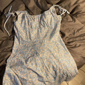 Blommig klänning från Shein - Söt blommig klänning från Shein i storlek S. Den är ljusblå med små blommor i vitt och orange. Klänningen har knytband vid axlarna och är ärmlös, perfekt för sommardagar. Materialet känns lätt och luftigt, vilket gör den idealisk för varma väder.