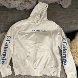 Vit hoodie från Pier One - Säljer en snygg vit hoodie från Pier One med blå text 'Colorado' på bröstet och ärmarna. Den har en klassisk känguruficka och en bekväm huva med dragsko. Perfekt för en avslappnad stil eller en kylig dag. Passar både till vardags och chillkvällar. 🏔️