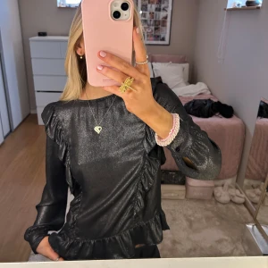 Svart glittrig blus med volangdetaljer - Säljer en superfin svart blus med glittrigt material och volangdetaljer. Perfekt för festliga tillfällen! Blusen har långa ärmar och en bekväm passform. Den är både stilren och trendig, och passar perfekt till både jeans och kjol. 🖤✨