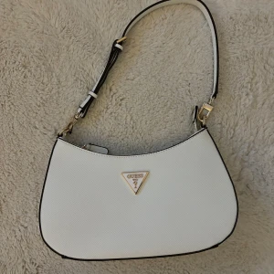 Vit axelväska från Guess - Säljer en snygg vit axelväska från Guess i mycket bra skick. Väskan har en stilren design med en dragkedja och justerbar axelrem. Perfekt för både vardag och fest! 👜✨