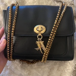 Svart axelväska med guldkedja - Snygg svart axelväska i skinn med guldkedja och coola detaljer som en dinosaurie, fjäder och stjärna. Väskan har ett rött innerfoder och praktiska fack med dragkedja. Perfekt för både vardag och fest!