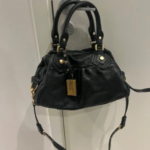 Marc Jacobs väska  - Säljer en snygg svart handväska i skinn med gulddetaljer. Väskan har både handtag och en justerbar axelrem. Insidan är fodrad med ett coolt mönster av bokstäver. Perfekt för både vardag och fest! 🎈Kan möta upp i centrala Stockholm 📍