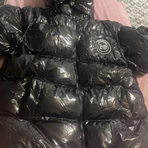 Svart glansig pufferjacka - Säljer en svart glansig pufferjacka från XO. Den är perfekt för kalla dagar med sitt quiltade mönster och långa ärmar. Jackan har en dragkedja framtill och en mysig huva. Den är i mycket bra skick och redo för vinterns äventyr! 🖤