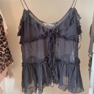 Slutsåld Zara topp - Säljer en superfin grå ärmlös topp från Zara i ett lätt och luftigt material. Toppen har volangdetaljer och knytning framtill, perfekt för sommardagar eller en festlig kväll. Den är i nytt skick och redo att bäras med stil!