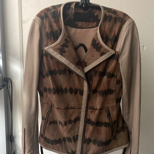 Mönstrad skinnjacka från Frontrow - Säljer en unik skinnjacka från Frontrow i bruna och beige toner med ett coolt mönster. Jackan har långa ärmar och dragkedja framtill. Perfekt för höst och vår när du vill sticka ut lite extra. Materialet är en mix av äkta mjuk  skinn och slipat läder
