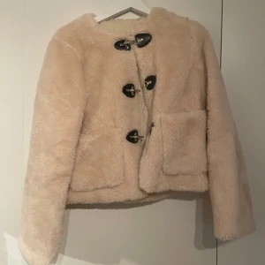 Beige teddyjacka med knappar - Supermysig beige teddyjacka med stora svarta knappar framtill. Perfekt för kyliga dagar och ger en stilren look. Jackan har långa ärmar och två stora fickor. Den är både varm och trendig, perfekt för höst och vinter.