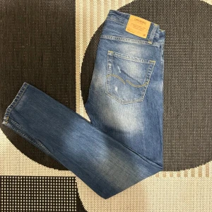 Blå jeans från Jack & Jones - Säljer ett par snygga blå jeans från Jack & Jones i storlek 30/32. De har en cool slitningseffekt och är i bra skick. Perfekta för en casual look med en t-shirt eller hoodie. Passar både till vardags och fest!