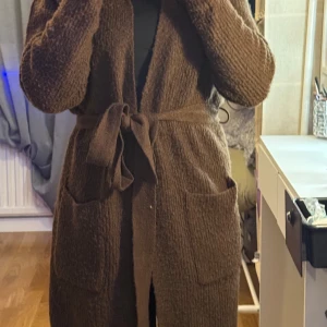Brun stickad kappa - Mysig brun stickad kappa med knytband i midjan. Perfekt för höst och vinter, den har långa ärmar och en skön, varm känsla. Passar bra för en avslappnad stil och håller dig varm under kyliga dagar.