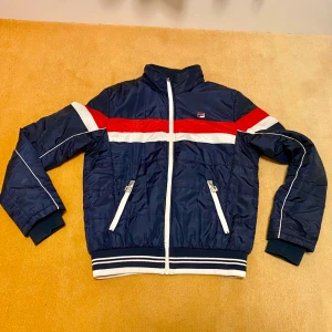 Blå täckjacka från Fila - Säljer en snygg retro blå jacka från Fila med vita och röda detaljer. Jackan har en dragkedja framtill och två fickor med dragkedjor. Perfekt för höst och vinter, med en retro look och bekväm passform. Den är i mycket bra skick och redo för nya äventyr! Storlek S - se bilder för mått.