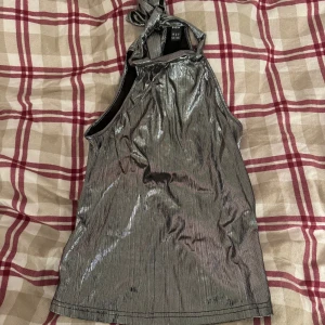 Glittrig silverfärgad halterneck-top - Snygg glittrig halterneck-top i silverfärg, perfekt för festliga tillfällen. Toppen har en knytning i nacken och är ärmlös, vilket ger en elegant look. Materialet är glansigt och fångar ljuset på ett coolt sätt. Perfekt för en utekväll eller nyårsfest!