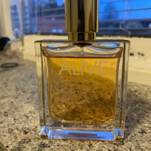 Parfym Alive - Säljer en elegant parfymflaska från Hugo Boss med namnet Alive. Flaskan är rektangulär och genomskinlig med en gyllene vätska inuti. Den har en lyxig guldfärgad kork. Doften har toppnoter av äpple, svarta vinbär, plommon, kanel och vanilj, hjärtnoter av jasmin och timjan, samt basnoter av cederträ, sandelträ, olivträ och nutkacypress. Flaskan innehåller 50ml 