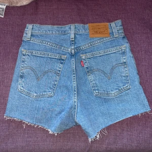 Blå jeansshorts från Levi's - Säljer ett par snygga blå jeansshorts från Levi's i storlek 25. De är högmidjade med en knappgylf och har en rå kant nertill för en cool look. Perfekta för sommaren och festivaler. Dom är i nyskick! Jag köpte dom för 1000kr.