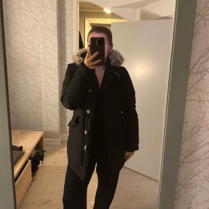 Svart parkajacka med päls från Woolrich - Säljer en svart parkajacka från Woolrich i mycket bra skick. Jackan har en mysig pälsfodrad huva och stängs med både dragkedja och knappar. Perfekt för kalla vinterdagar! 🖤